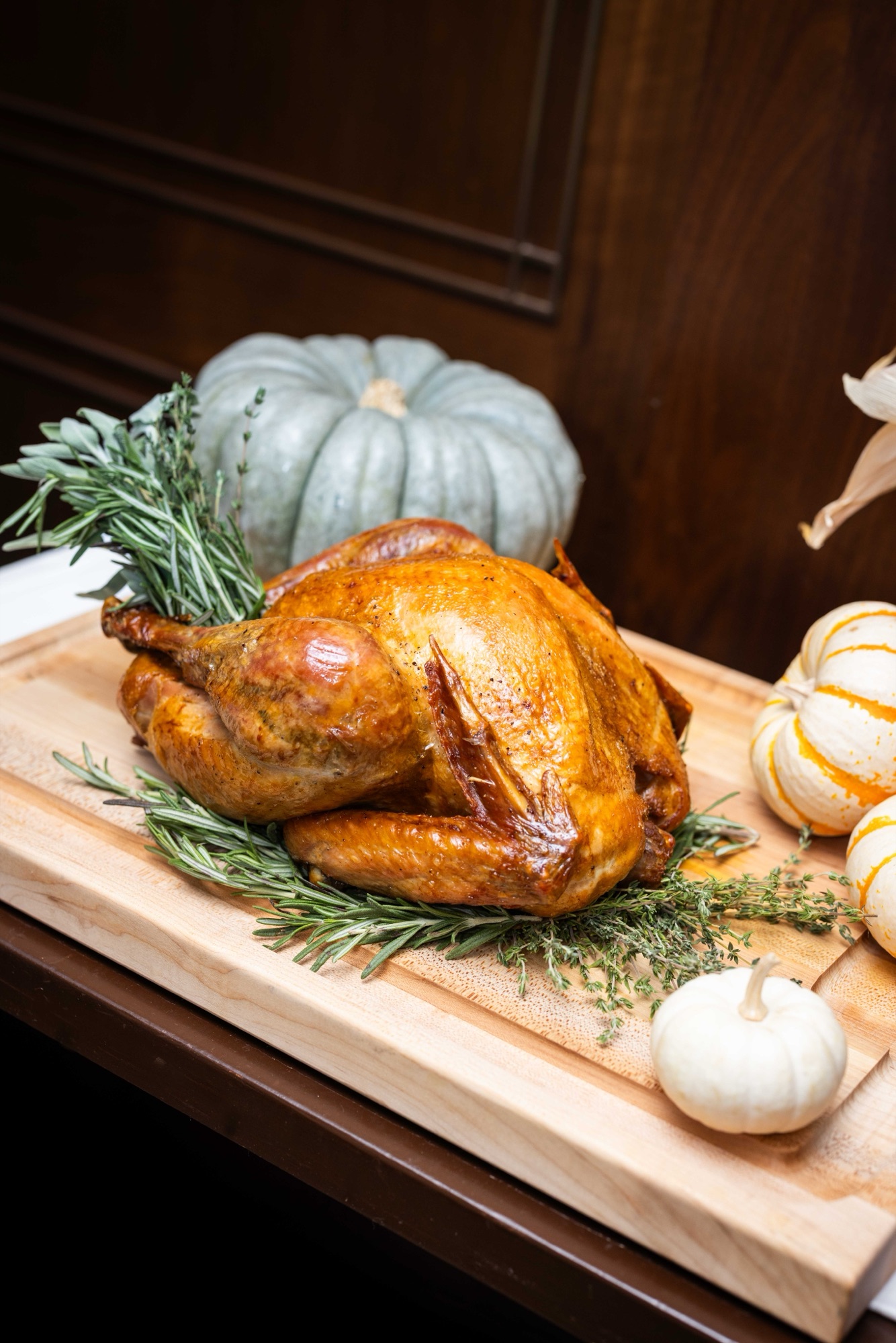 Whole roast turkey on a carving board with rosemary, surrounded by white and blue-gray heirloom pumpkins