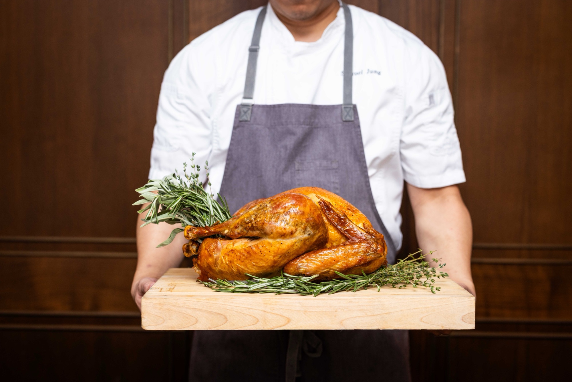 A chef holds a golden roast turkey on a wooden carving board garnished with fresh rosemary